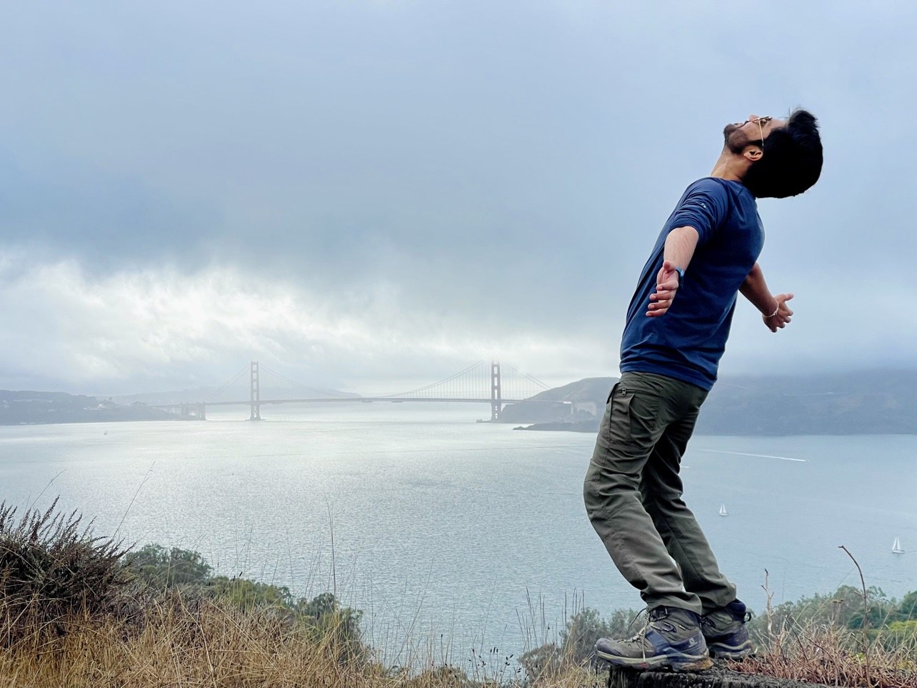 Sagar leaning back with arms outstretched, the Golden Gate Bridge fading into fog across the bay behind him, seen from Angel Island
